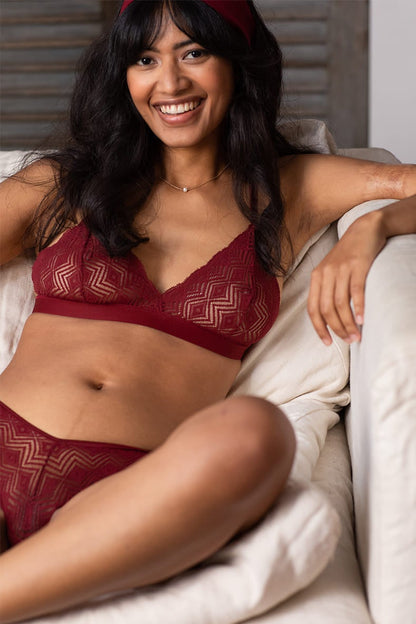 Jeune femme brune et souriante porte un ensemble de lingerie rouge foncé confectionné en coton bio et en dentelle recyclée.