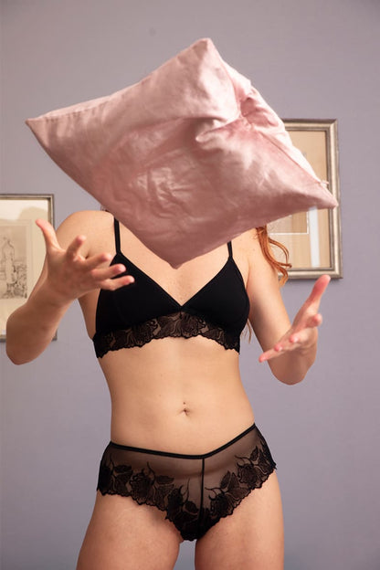 Femme en lingerie de la marque Olly, jouant avec un coussin en velours rose, elle porte un ensemble de lingerie éco-responsable, dont un shorty et soutien-gorge noir en dentelle recyclée.